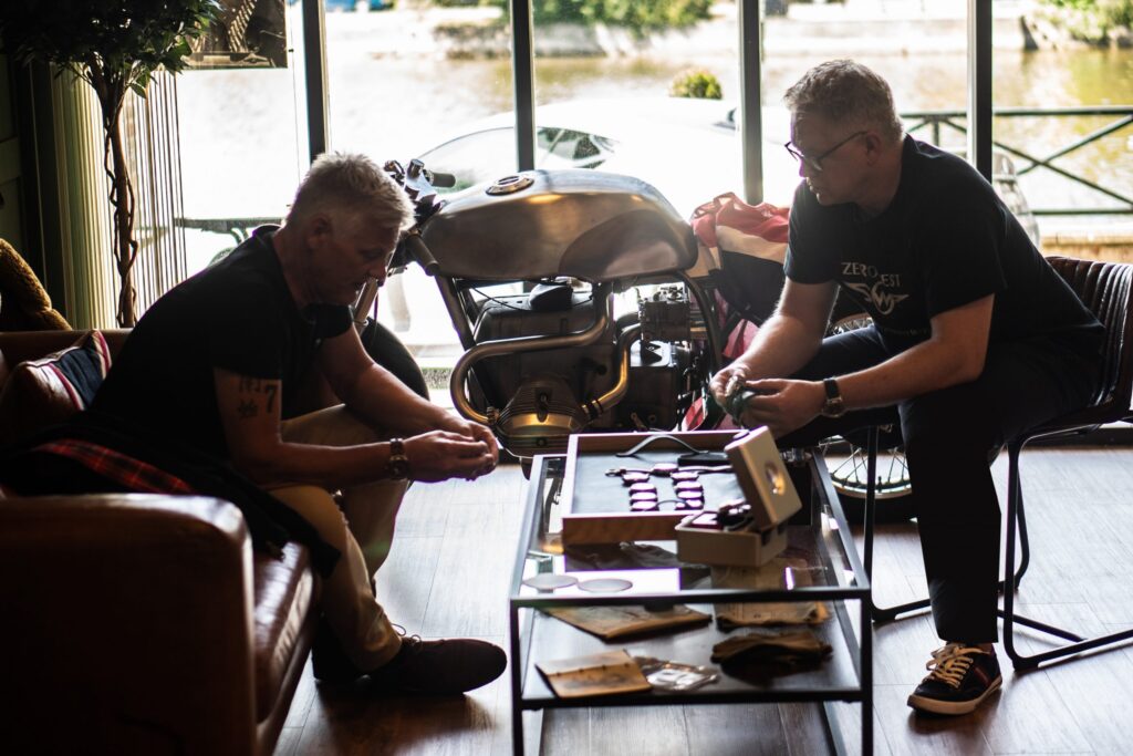 Two men sitting next to a motorbike looking at watches